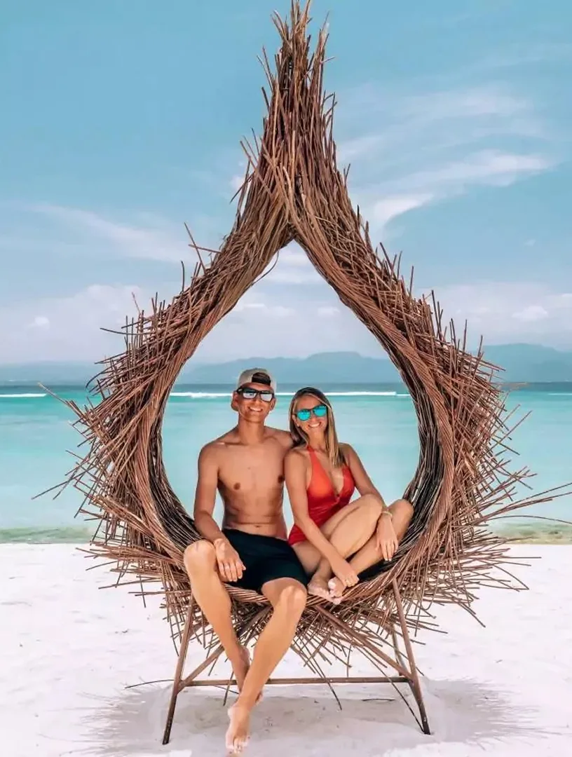 Daniel und Jessi sitzen gemeinsam in einem geflochtenen Strandkorb am weißen Sandstrand mit türkisblauem Meer im Hintergrund