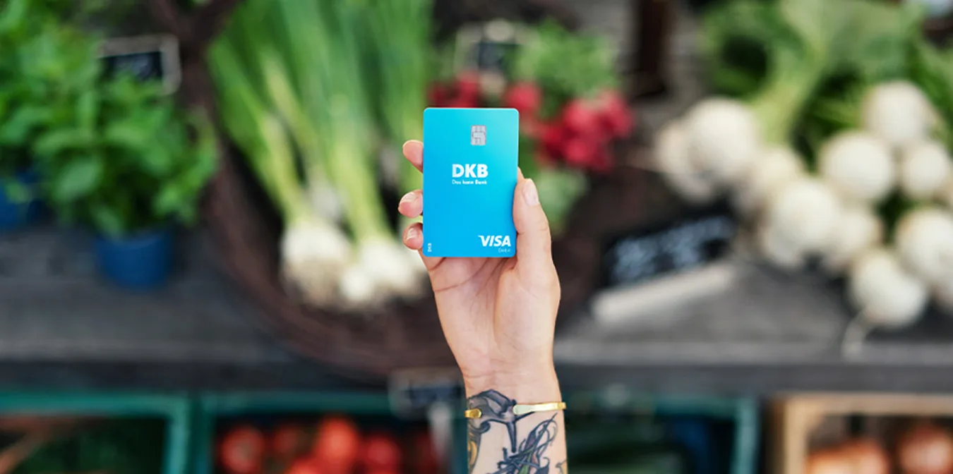 Hand hält DKB Visa Karte vor unscharfem Hintergrund auf einem Marktstand mit Gemüse.