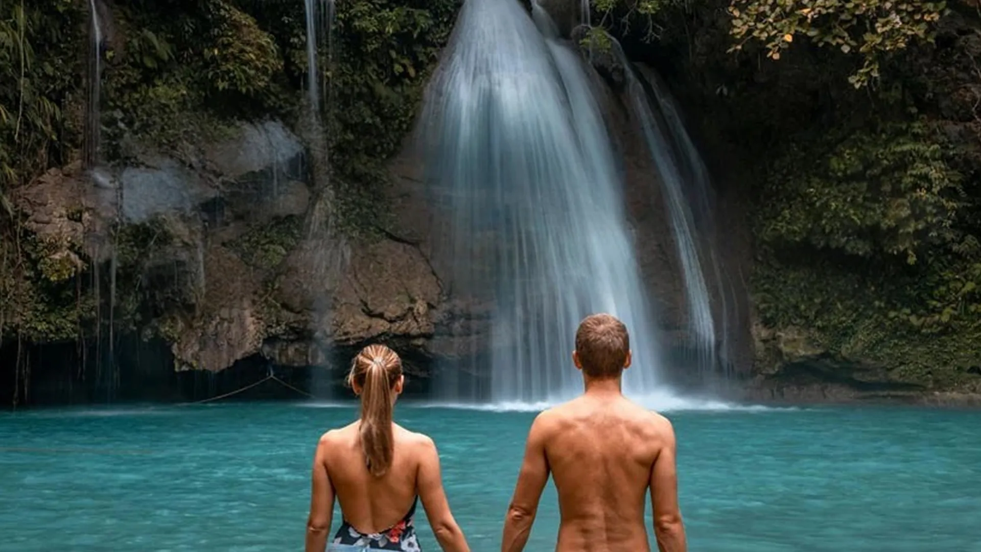 Paar steht vor Wasserfall in türkisfarbenem See in tropischer Umgebung.